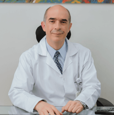 Smiling bald doctor in white lab coat and tie, seated at a desk.