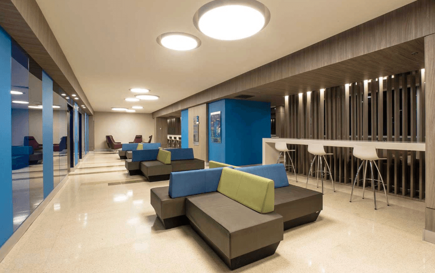 Bright modern lounge with blue and green modular sofas, circular lights, and a white bar.