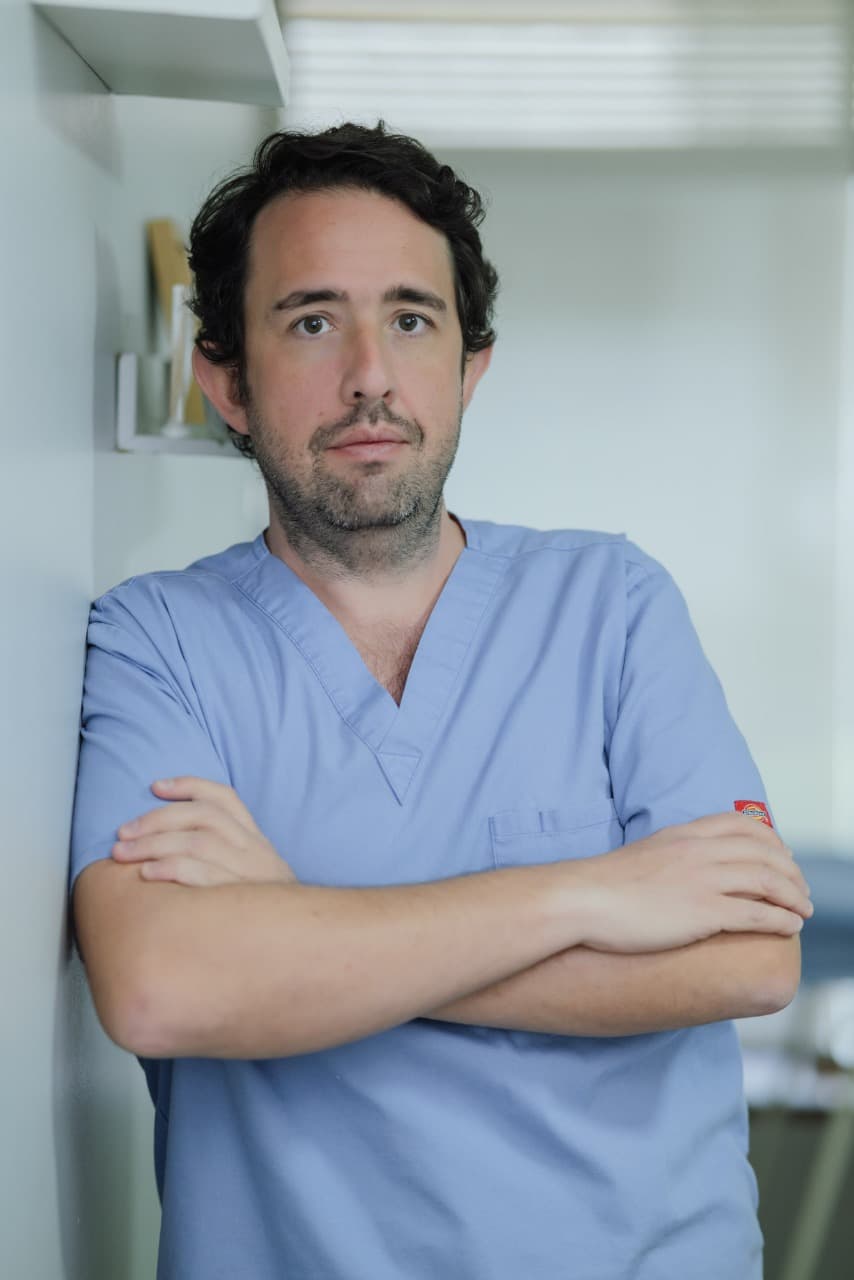 Dark-haired man in light blue scrubs, arms crossed, leaning against a white wall.